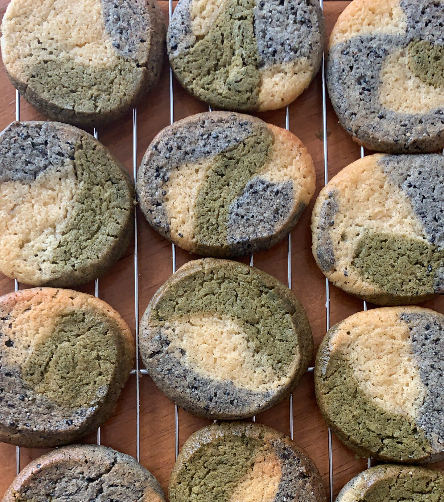 Photo of 12 cookies with a marble pattern of green (matcha), black sesame (black) and vanilla (white) on a baking rack.